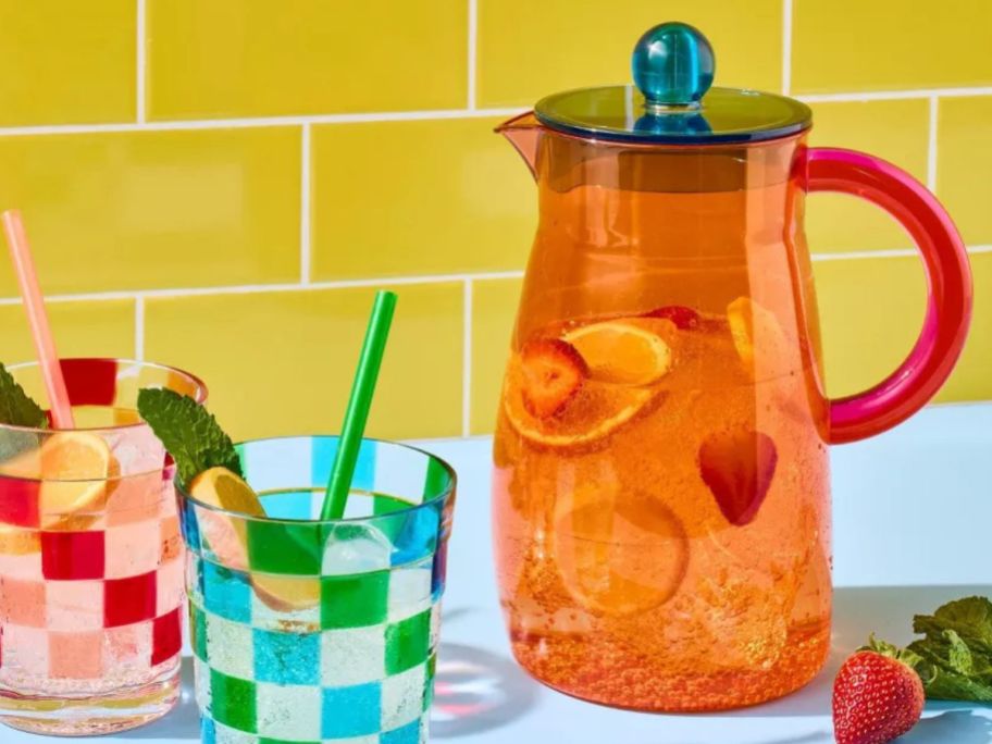 orange and blue Tabitha Brown 64oz Plastic Beverage Pitcher with ice and fruit in it next to tumblers on counter